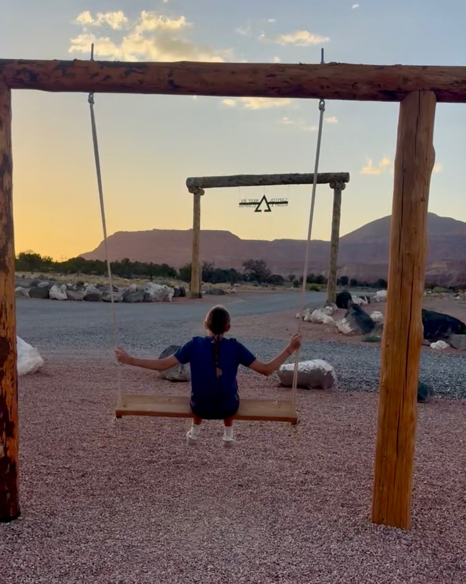 Child on a log swing facing the resort sign and red-rock landscape at sunset