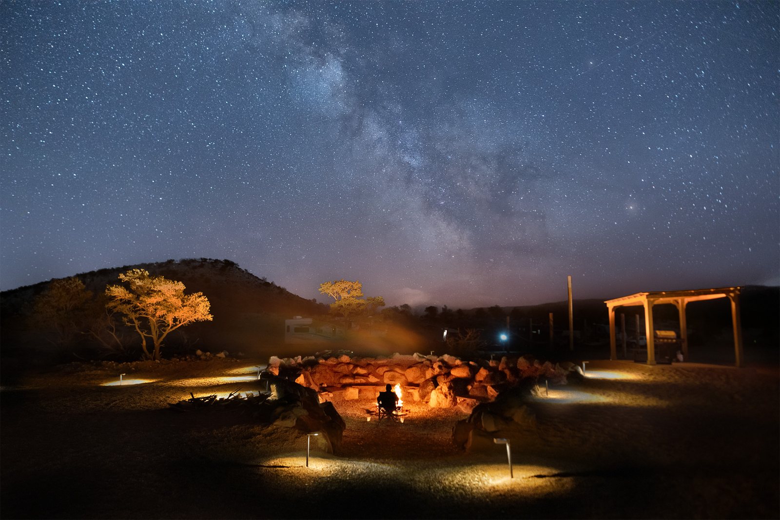 A guest seated at the resort fire pit beneath the Milky Way, with low pathway lights nearby
